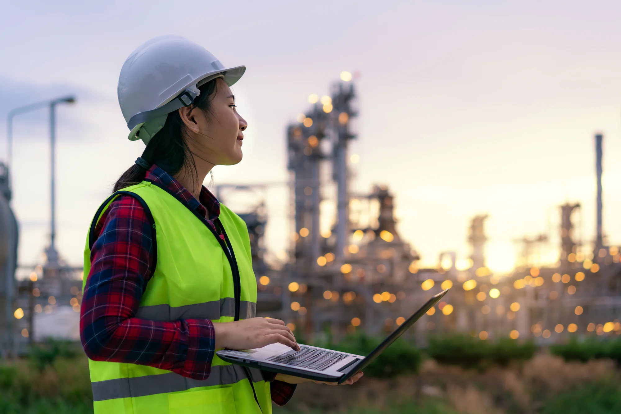 A worker in a hard hat and green reflecting vest looks at an industrial power plant while holding a laptop.