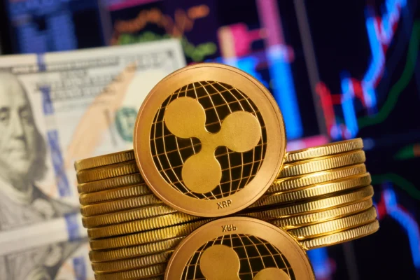 Gold coin with the Ripple/XRP logo, in front of more coins and $100 bills.