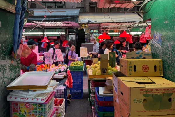 A bustling fruit market with various stalls selling produce. People are gathered around the stands displaying fruits like oranges and mangoes. The market is lit by red pendant lights and has cardboard boxes stacked in the foreground. Some signs are visible, written in a language with Chinese characters.