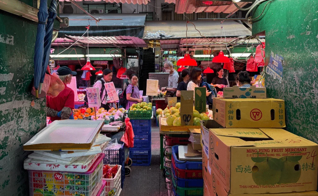 A bustling fruit market with various stalls selling produce. People are gathered around the stands displaying fruits like oranges and mangoes. The market is lit by red pendant lights and has cardboard boxes stacked in the foreground. Some signs are visible, written in a language with Chinese characters.