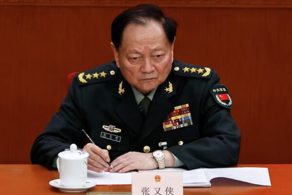 A man with black hair, wearing a military uniform, holds a pen as he sits at a desk.