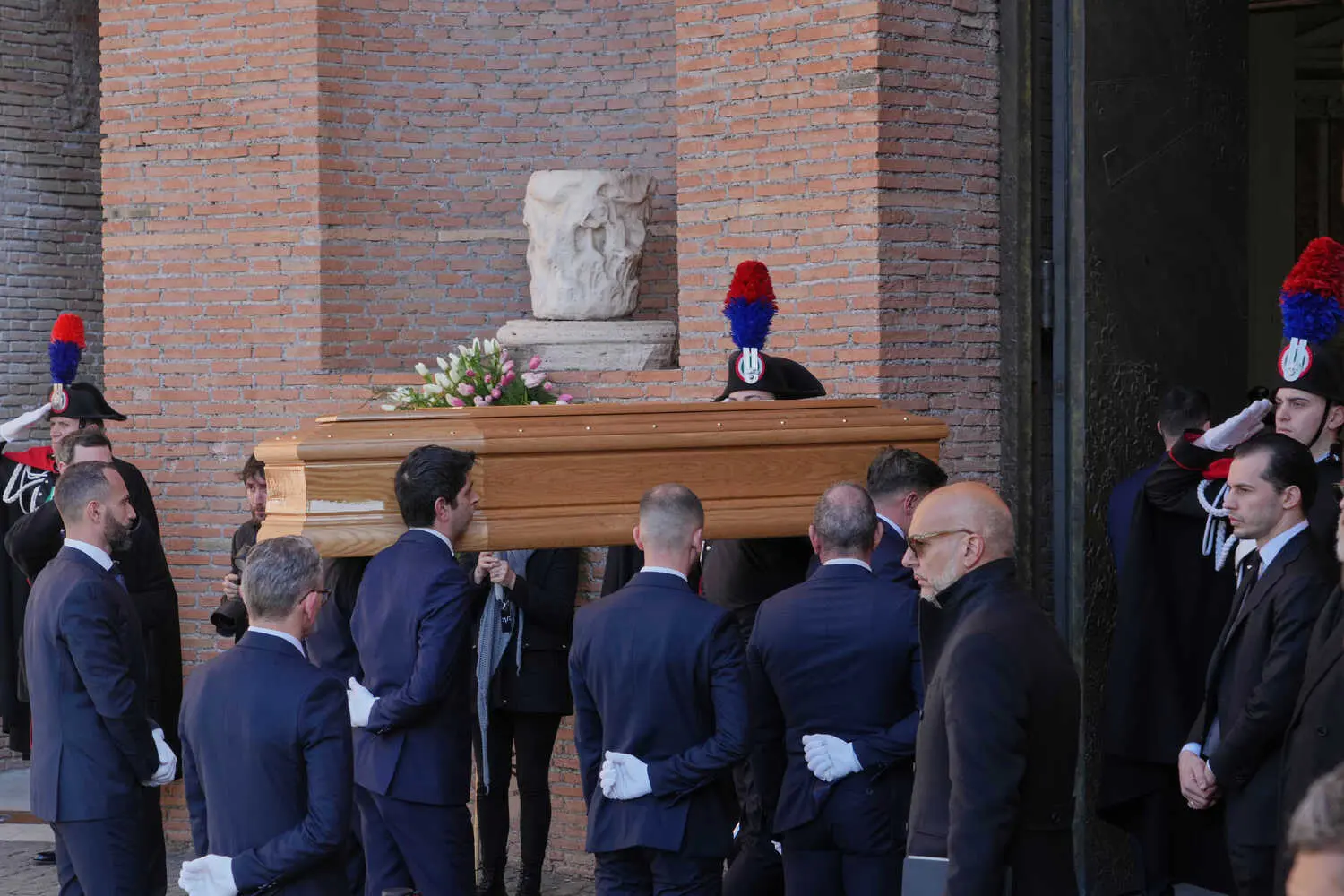A wooden casket with white and pink flowers is carried by people in dark suits. A brick wall and uniformed people with red and blue hats are in the background.
