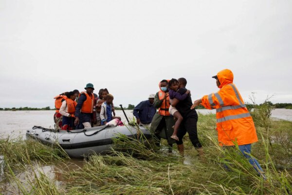 A few adults, one carrying a child, wade through flooded vegetation toward an inflatable boat that already has several people in it.