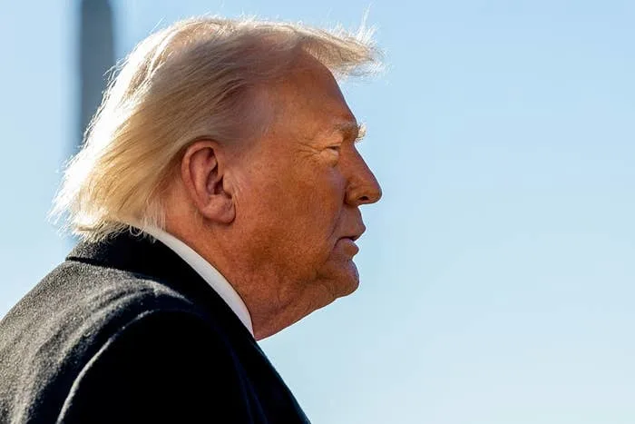 A person in side profile with windswept hair, wearing a suit, appears outdoors with a blurred background