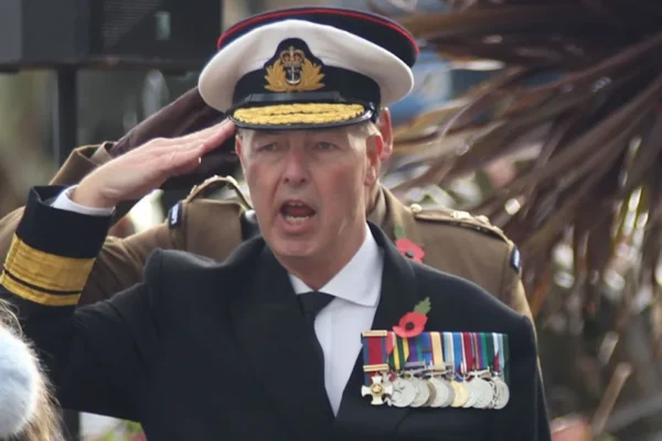 A man dressed as an admiral gives a salute. He wears a white hat, navy jacket, white shirt and tie. On his chest are a series of medals hung by colourful ribbons.