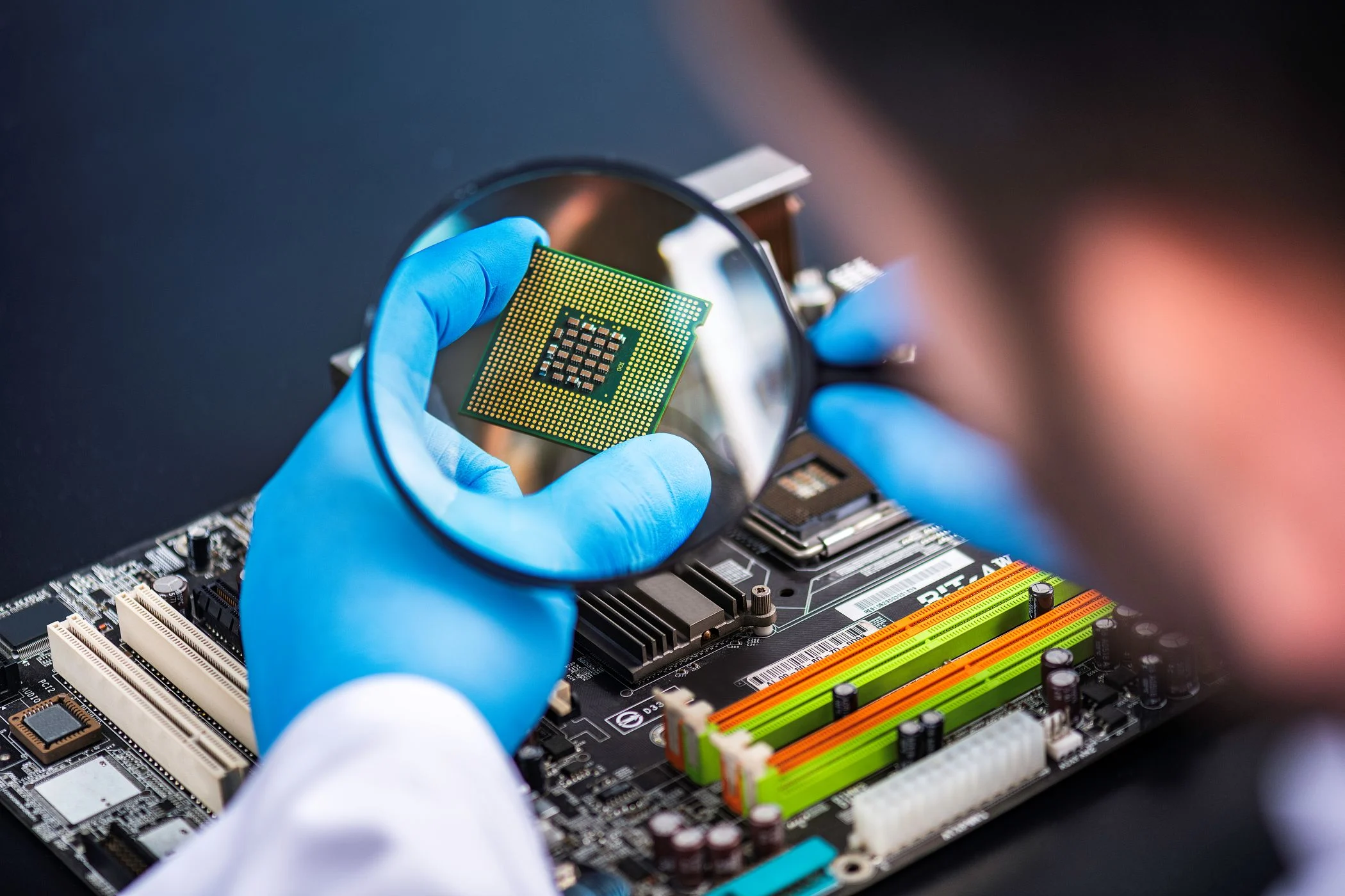 Blue-gloved hands examine a semiconductor chip under a magnifying glass.