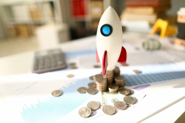 A toy rocket ready for launch atop messy stacks of coins and paperwork displaying financial data.