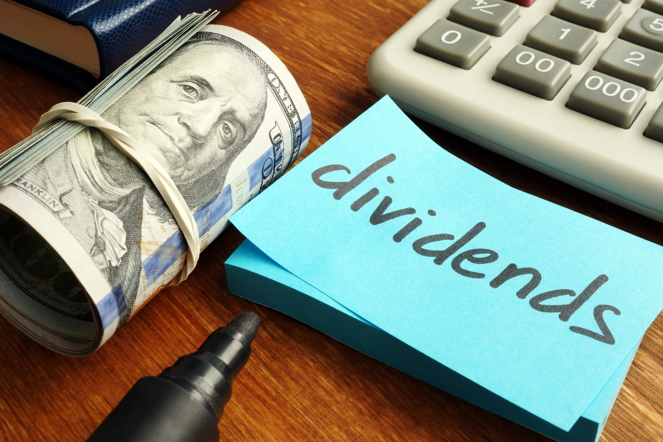 A roll of hundred-dollar bills is next to a post-it with the word dividends written on it.
