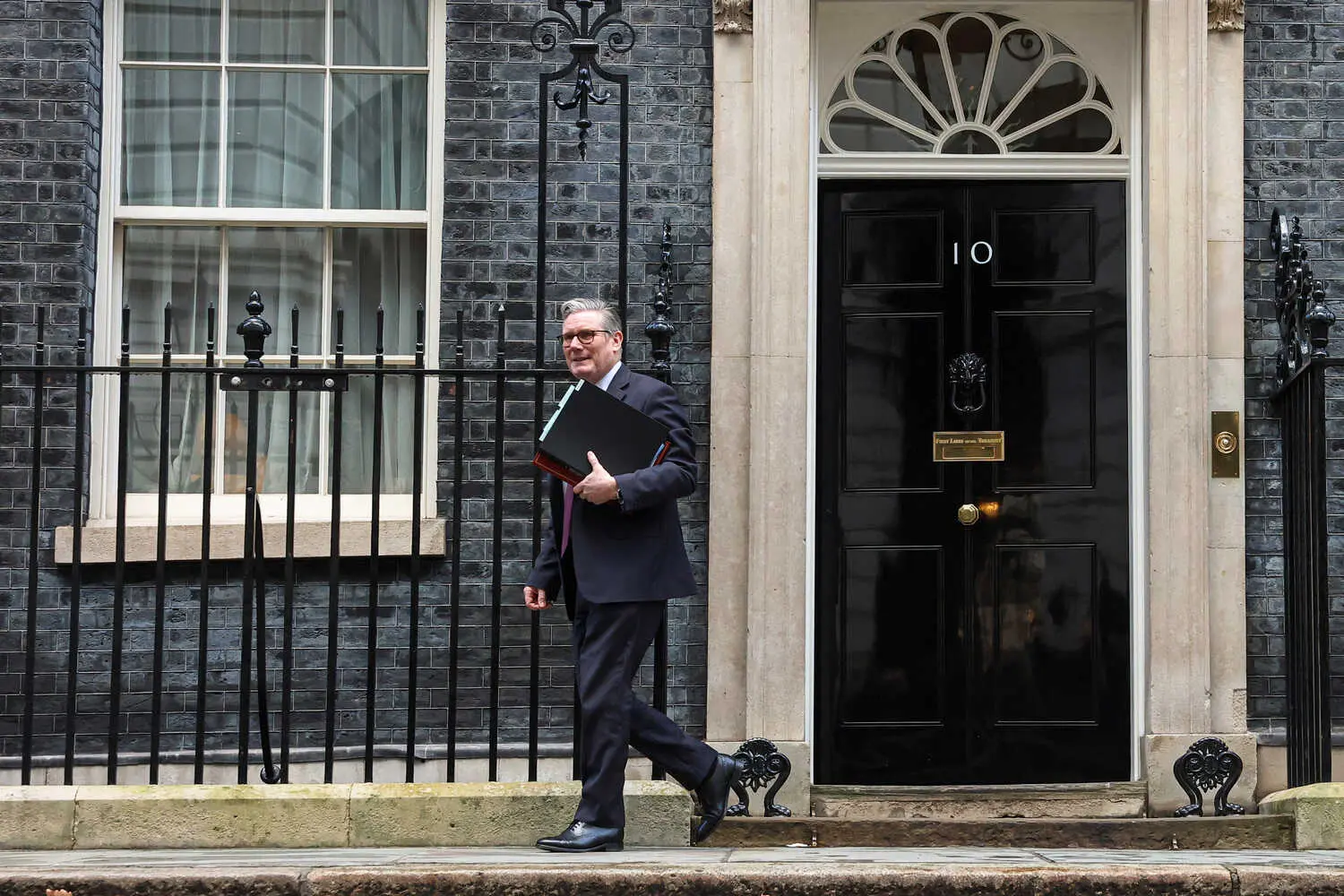 Keir Starmer, in a dark suit, carries a stack of folders as he walks in front of the black door of No. 10 Downing Street.
