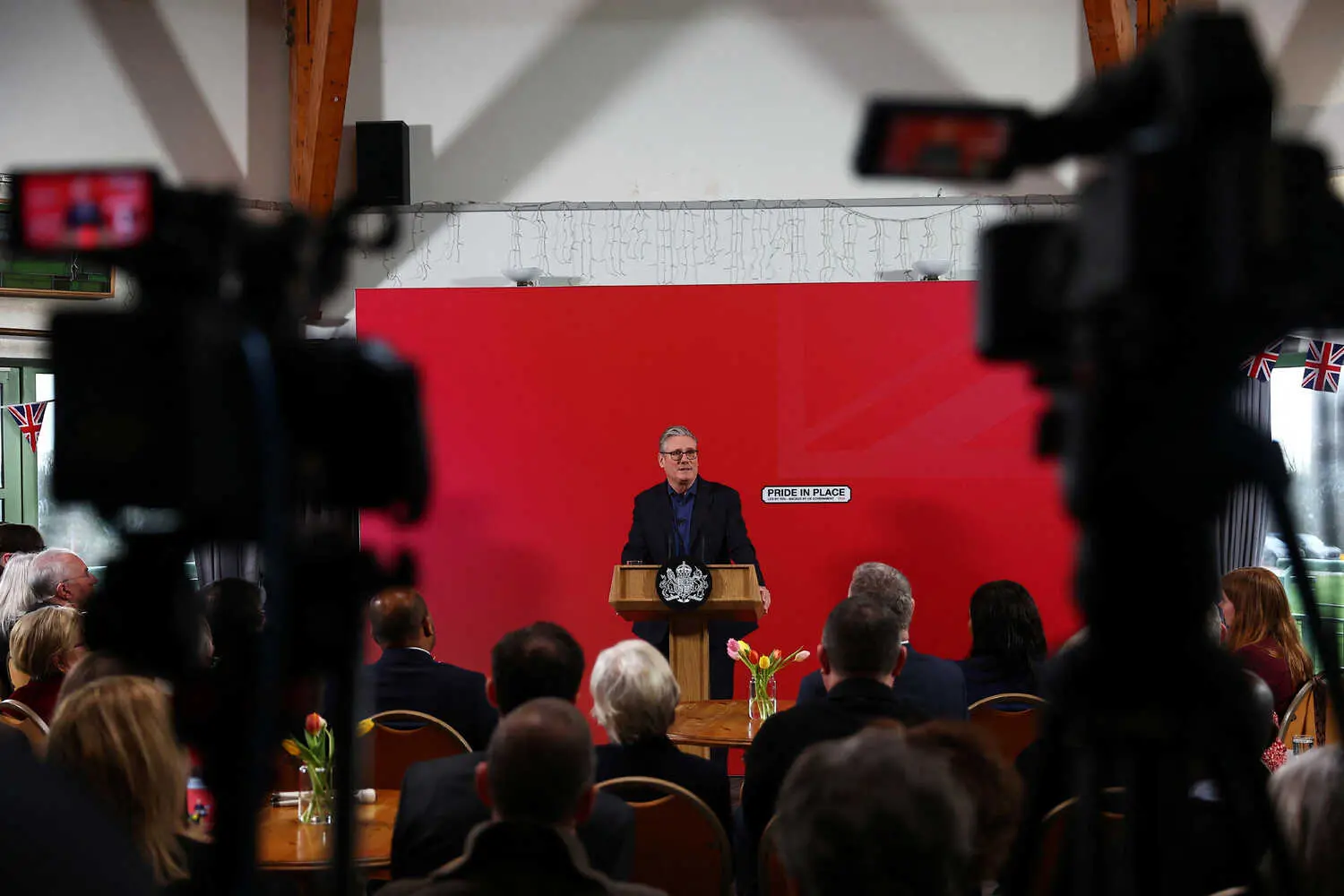 Keir Starmer speaks at a lectern, facing an audience. Behind him is a red wall with "PRIDE IN PLACE" and Union Jack flags.