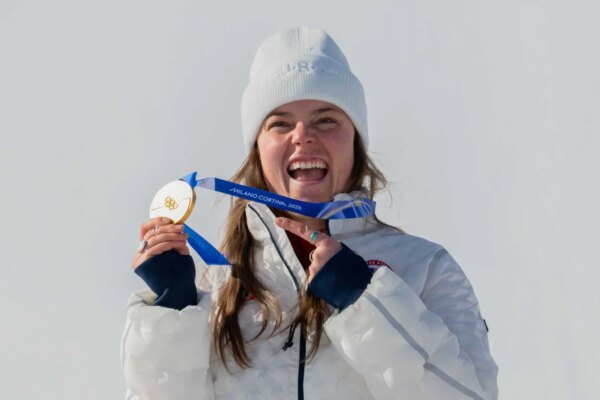 Breezy Johnson, wearing a white beanie hat and a white coat, smiling and holding up her gold medal.