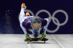A man holding a skeleton sled on an ice track, wearing a helmet with images of people on it.