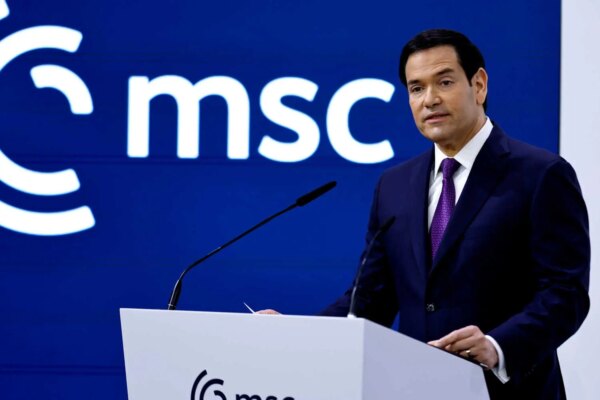 Marco Rubio, wearing a dark suit and a purple tie, stands at a white lectern with two microphones. A blue wall with a white “msc” logo is behind him.
