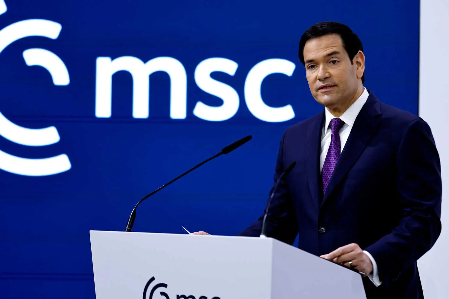 Marco Rubio, wearing a dark suit and a purple tie, stands at a white lectern with two microphones. A blue wall with a white “msc” logo is behind him.
