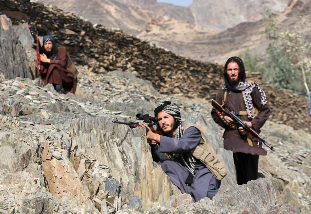 Three people with rifles are positioned on a rocky mountain slope. One person in the foreground crouches, aiming a rifle with a scope.