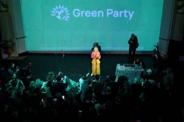 A woman dressed in yellow and pink speaks underneath a large screen with a Green Party emblem projected on it.