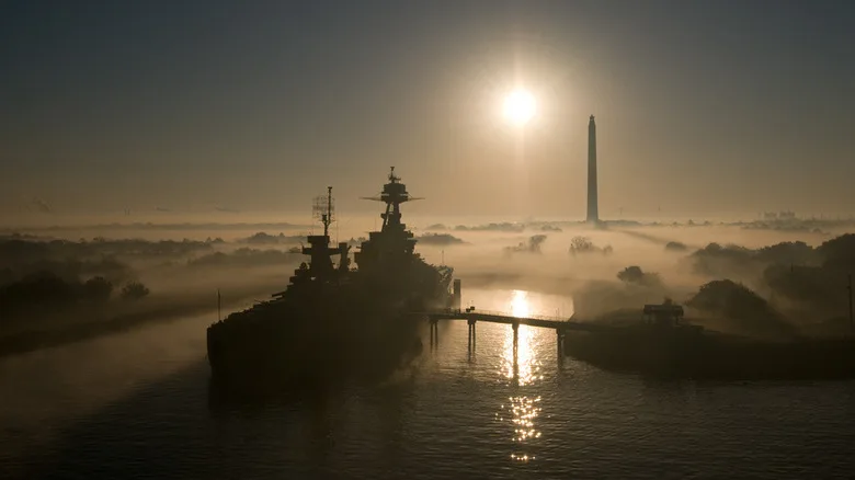 Battleship USS Texas seen from a high distance front port side view in morning fog with San Jacinto Monument in background