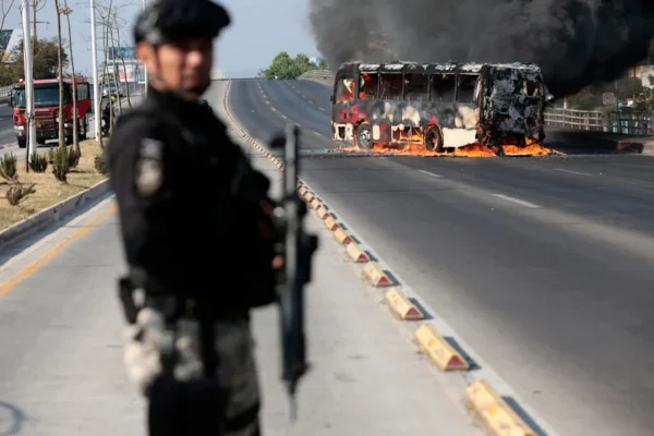 A member of the Prosecutor's Office stands guard near a burning bus  set on fire by organized crime groups in response to an operation in Jalisco to arrest a high-priority security target, at one of the main avenues in Zapopan, state of Jalisco, Mexico, on Feb. 22, 2026. / Credit: Ulises Ruiz /AFP via Getty Images