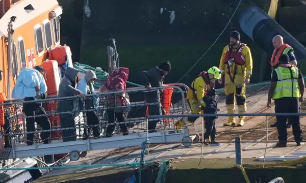 People walking a gangway from the ship to the harbour. Many wear blankets around them. Four emergency personnel stand on the harbour waiting for the migrants. One child is being helped by the worker