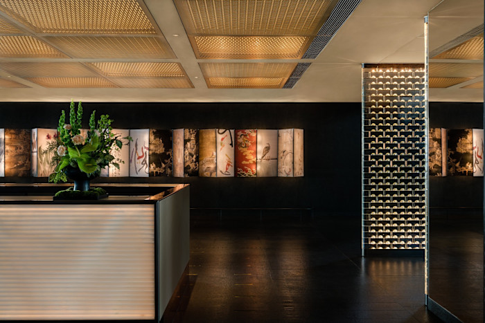 A long, illuminated art montage with panels of traditional and modern motifs is displayed on a dark wall behind a reception desk with a large green floral arrangement in the Regent Hong Kong.
