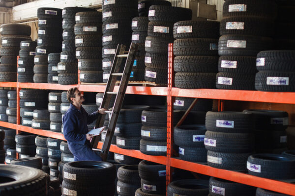 Mechanic holding a new car tire looking upward to shelf of tire inventory