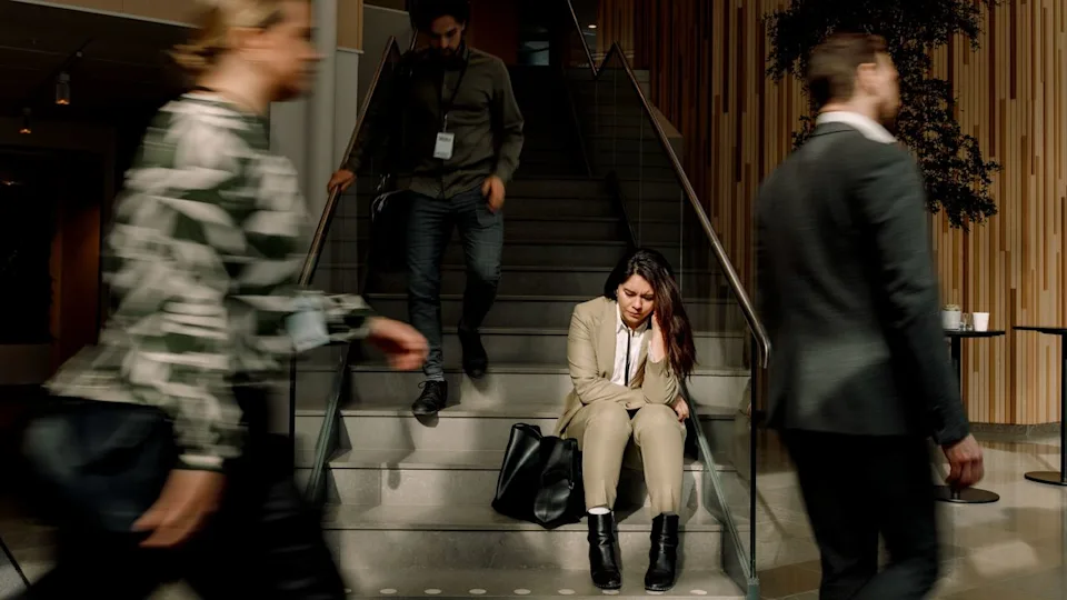 Frustrated woman in a business suit sitting on stairs while other workers walk around her in a blur .