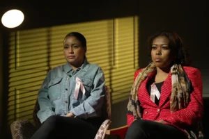 Elise Skillen and Savannah Holm-Aderemi are sitting beside each other, in a panelled room, with a yellow light shining against the wall. Skillen is wearing a denim jacket and Holm-Aderemi has a red jacket and a patterned scarf around her neck. Both are wearing pink ribbons in tribute to Fiona Holm