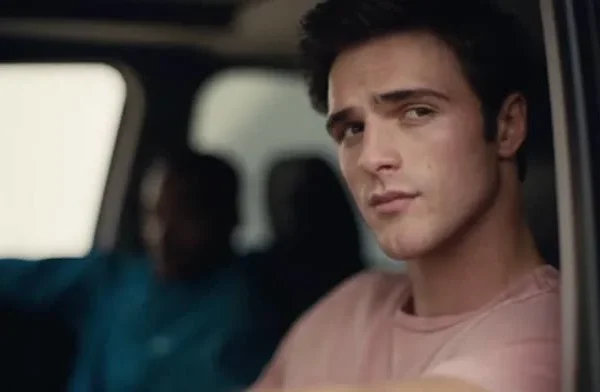 Person in a car looking out the window pensively. Another person is in the driver's seat