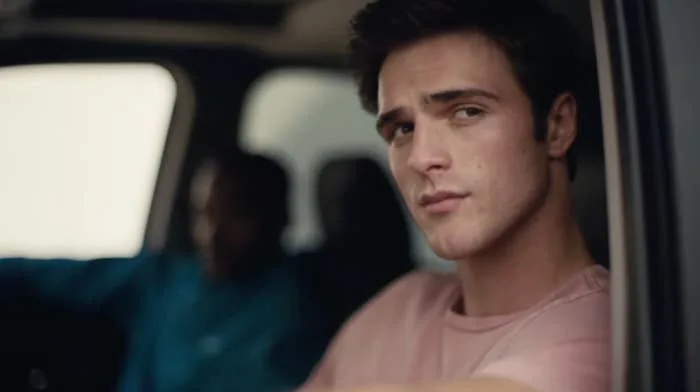 Person in a car looking out the window pensively. Another person is in the driver's seat