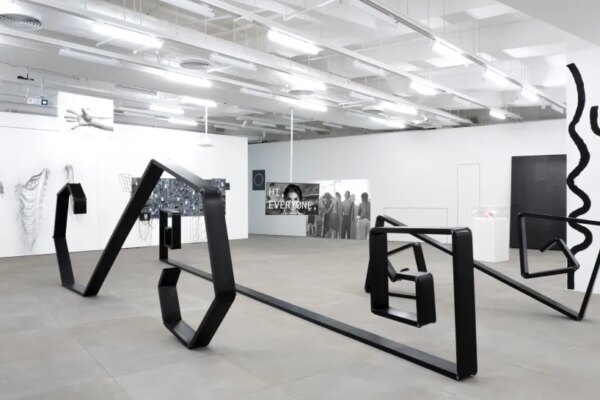 A spacious white gallery with industrial ceiling lights presents angular black sculptural forms arranged across the floor and a mirrored surface, surrounded by monochrome drawings, text-based works and wall-mounted pieces.