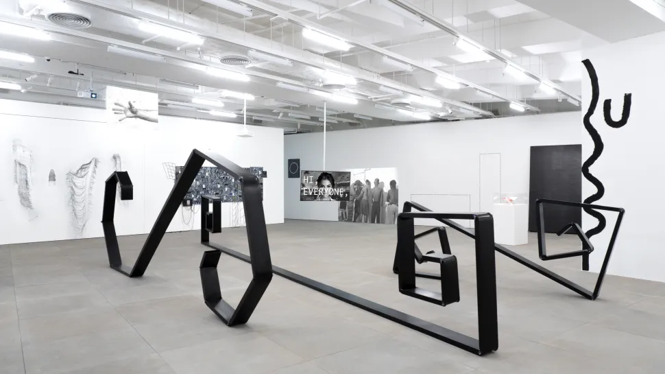 A spacious white gallery with industrial ceiling lights presents angular black sculptural forms arranged across the floor and a mirrored surface, surrounded by monochrome drawings, text-based works and wall-mounted pieces.