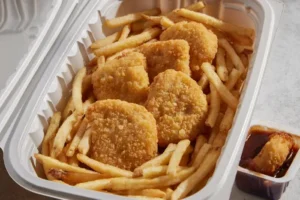 Chicken nuggets and fries in a takeout container, with a small sauce cup on the side, appearing fresh and crispy