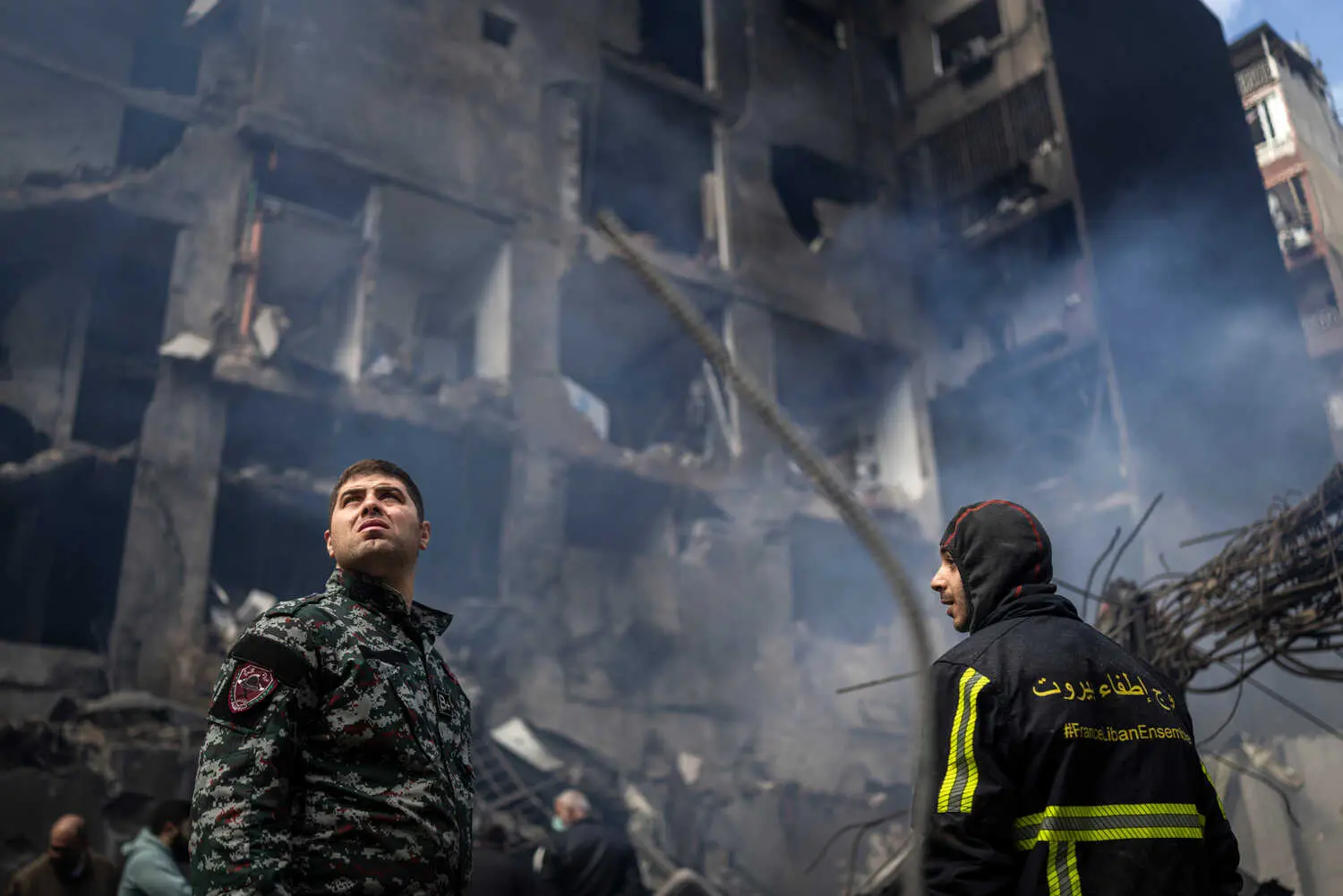 Two emergency workers look up at a damaged building with smoke. One wears a camouflage uniform; the other, a dark jacket with yellow stripes and text.