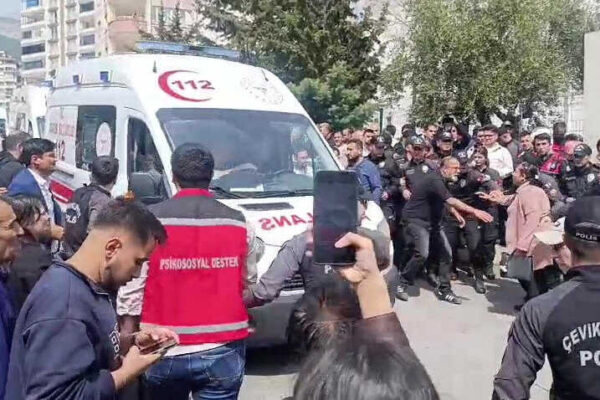 Numerous people, including many in police uniforms, gather around a white ambulance. Some individuals are seen in a confrontation.