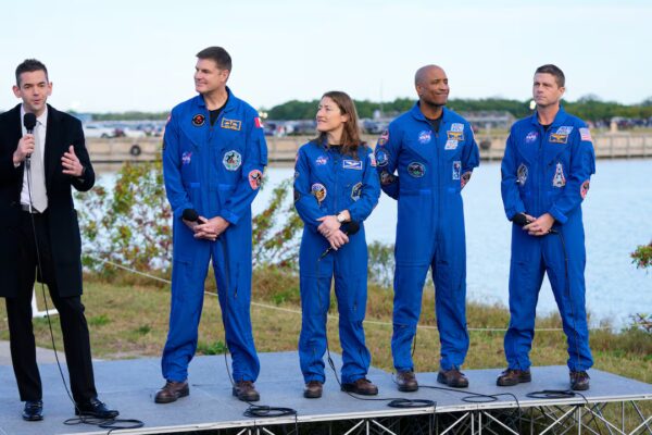 A man in a suit talks into a microphone next to four astronauts in blue space suits.