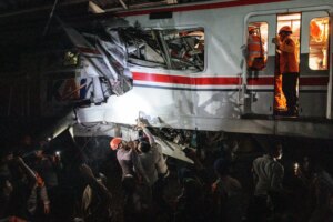 The mangled wreckage of a train. People gathered nearby are shining lights as rescue workers in orange helmets search the wreckage.