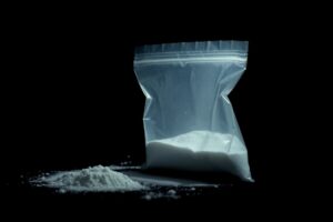 An extreme close-up photograph of a small plastic bag containing an unidentified white powder, lit by a harsh, direct camera flash against a pitch-black background, creating a stark, gritty, investigative aesthetic.
