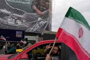 A man flashes a victory sign as he carries an Iranian flag in front of an anti-US billboard depicting the American aircrafts into the Iranian armed forces fishing net with signs that read in Farsi: "The Strait of Hormuz will remain closed, (AP)