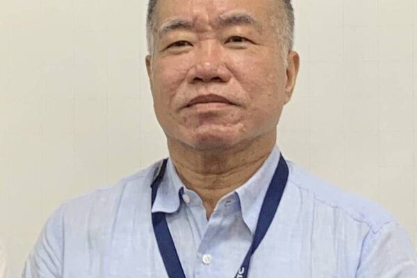 A person wearing a light blue shirt and a dark blue lanyard stands against a plain background.