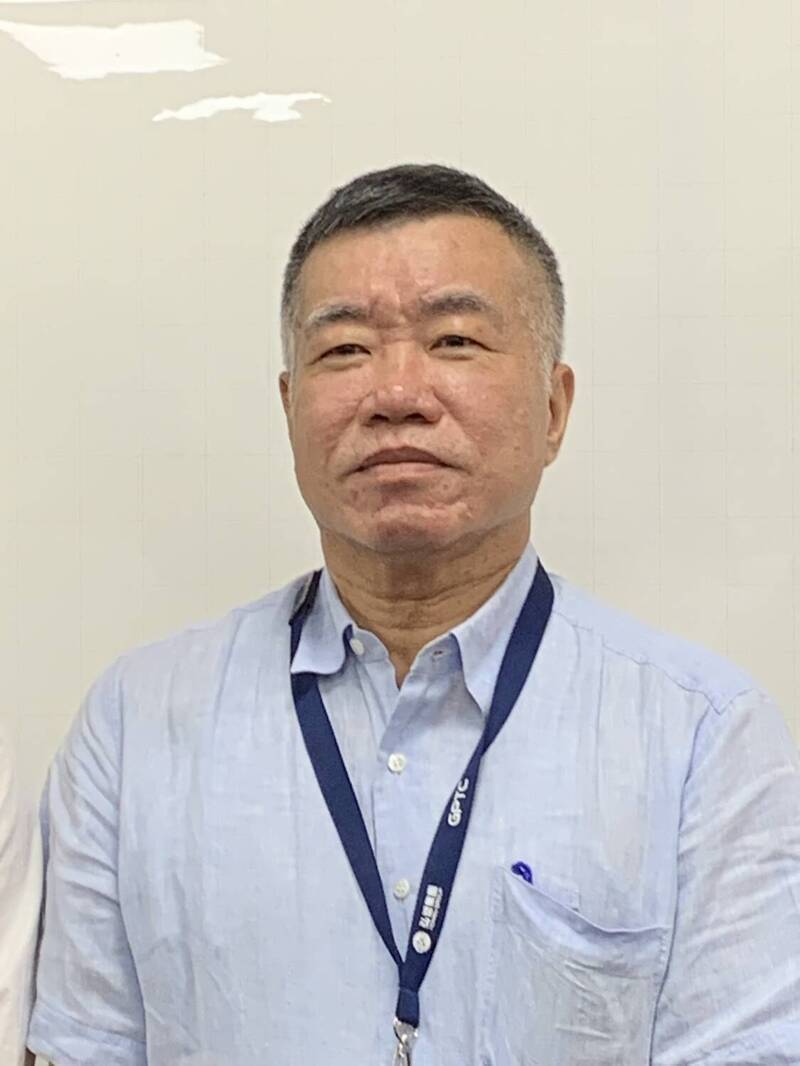 A person wearing a light blue shirt and a dark blue lanyard stands against a plain background.