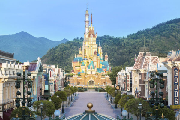Catch a glimpse of a colorful castle at the end of a bustling street lined with charming shops and lamp posts, with green mountains standing majestically in the background—a scene perfect for your daily recap.