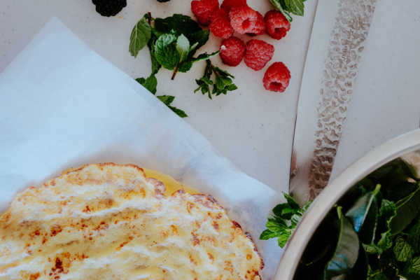 A golden, cooked flatbread rests on white parchment paper, surrounded by fresh raspberries, blackberries, mint leaves, a large textured knife, and a bowl of greens.