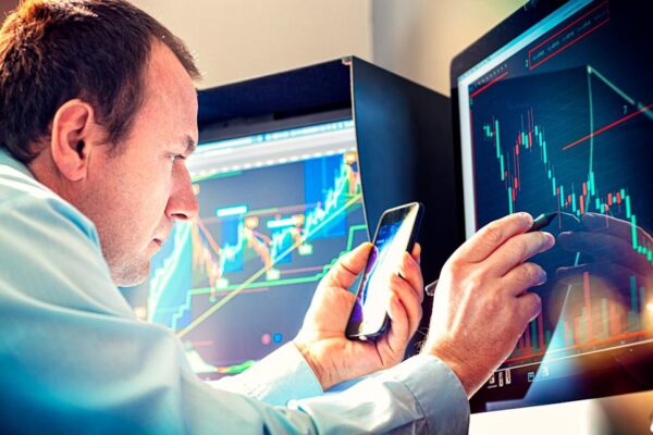 A money manager using a smartphone and stylus to analyze a stock chart displayed on a computer monitor.