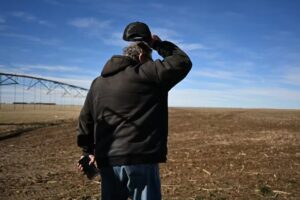 A man in a hoodie stands in a field, lifting his ballcap and scratching his head.