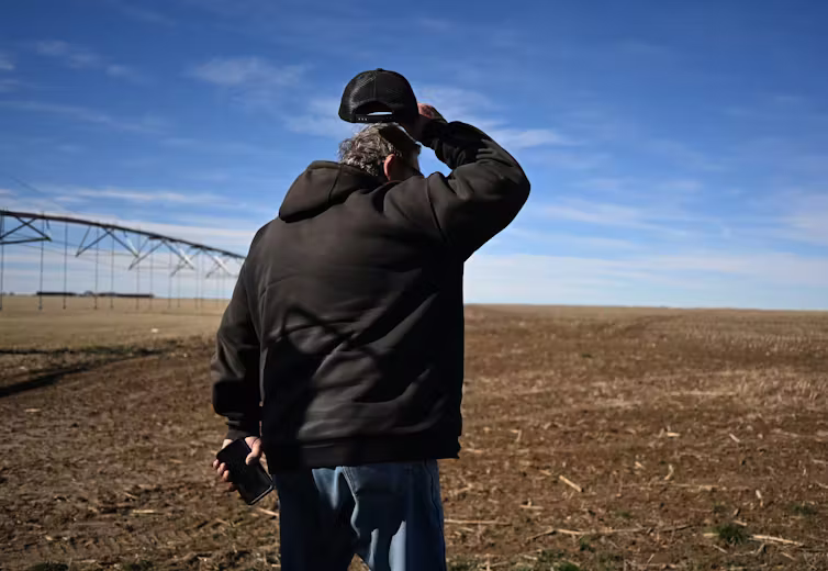 A man in a hoodie stands in a field, lifting his ballcap and scratching his head.