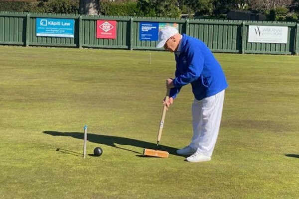 101-year-old Neville Sandiford in a white hat and blue jacket playing croquet on a green lawn.