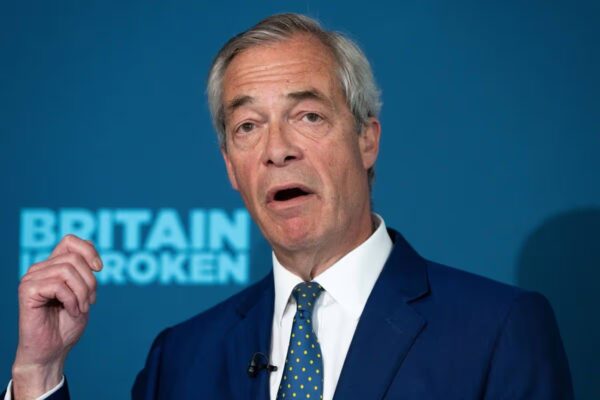 An individual, dressed formally, is engaged in a speech. The backdrop features a blue banner with the text "BRITAIN BROKEN" prominently displayed.