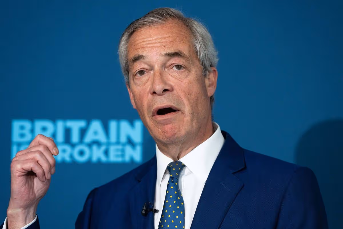 An individual, dressed formally, is engaged in a speech. The backdrop features a blue banner with the text "BRITAIN BROKEN" prominently displayed.