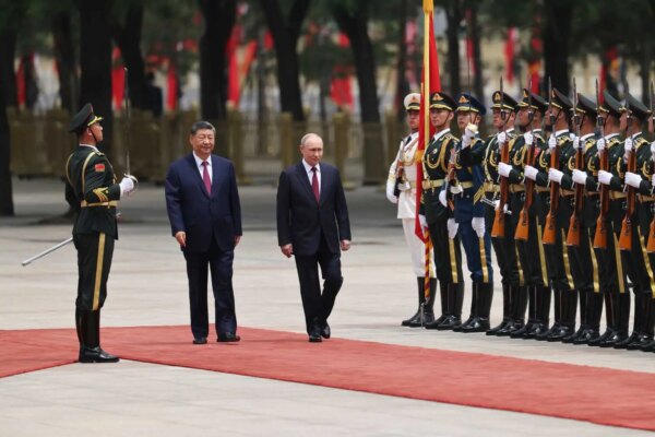 Xi Jinping and Vladimir Putin walk side by side on a red carpet. A line of soldiers stand to the side, holding guns.