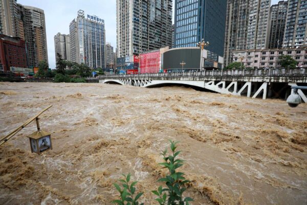Deep, brown water rushes under a bridge. Tall buildings are in the background.